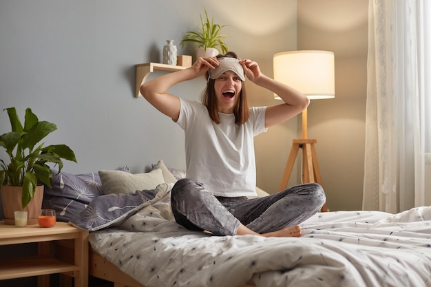 Afbeelding van opgewonden verbaasde vrouw met slaapmasker zittend op bed in knusse slaapkamer kijkend naar camera met aangename verrassing lachend gelukkig koel weekend