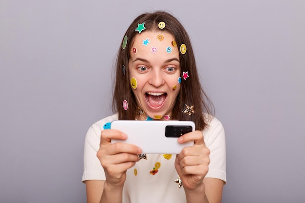 Afbeelding van opgewonden vrouw gamer bedekt met grappige stickers poseren geïsoleerd over grijze achtergrond spelen van videogames winnende niveau kijkend naar het scherm van de mobiele telefoon