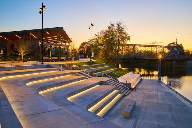 Afbeelding van park van cementtrappen op rivier met metalen loopbrug en zonsondergang