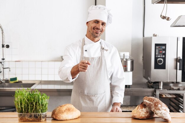 Afbeelding van positieve man bakker in wit uniform glimlachen, terwijl staande bij bakkerij met brood op tafel