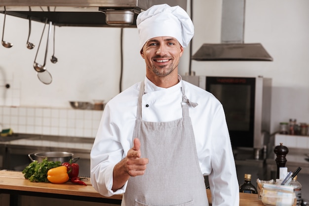 Afbeelding van positieve mannelijke chef in wit uniform glimlachen, terwijl staande op keuken in restaurant