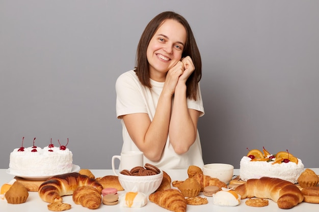 Afbeelding van schattige charmante schattige glimlachende vrouw met bruin haar zittend aan tafel tussen grote verscheidenheid aan bakkerijen kijkend naar camera geïsoleerd over grijze achtergrond