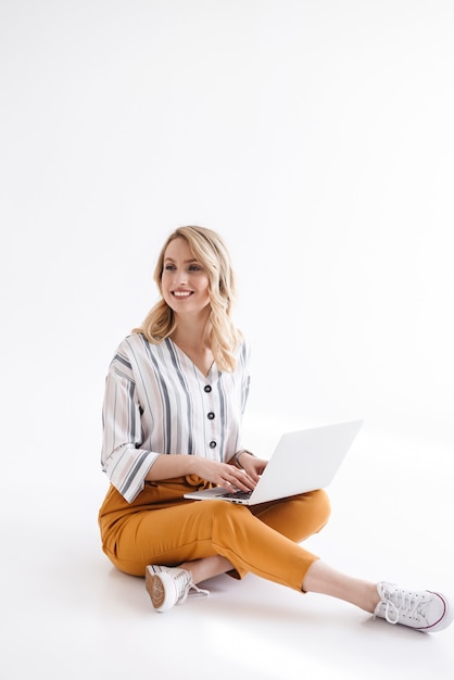 Afbeelding van schattige glimlachende vrouw die vrijetijdskleding draagt die op laptop typt en opzij kijkt terwijl zittend op de vloer die over witte muur wordt geïsoleerd
