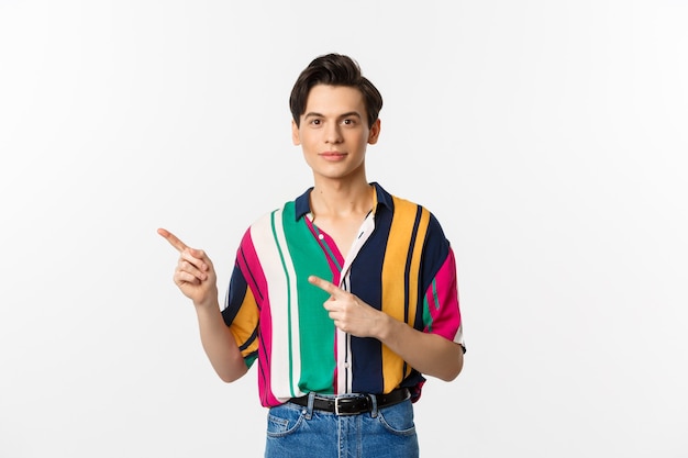 Afbeelding van stijlvolle androgyne man glimlachend, wijzende vingers links naar logo, promo-aanbieding, staande over wit