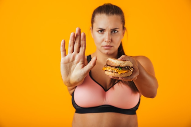 Afbeelding van strikte mollige vrouw in trainingspak doet stop gebaar terwijl sandwich, geïsoleerd op gele achtergrond