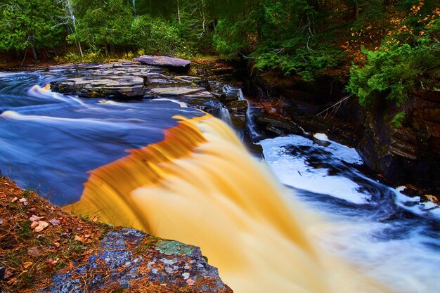 Afbeelding van stromend water in de rivier leidt naar een bruine en gouden waterval met een wervelend zwembad aan de basis in een rotsachtig gebied met dennennaalden en groen bos