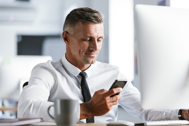 Afbeelding van succesvolle zakenman 30s dragen witte overhemd en stropdas zitten aan de balie in kantoor door computer, en mobiele telefoon te houden