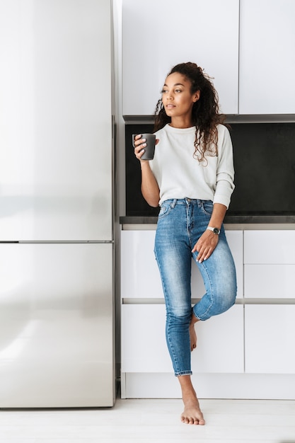 Afbeelding van tevreden Afrikaans Amerikaans meisje dat thee drinkt, terwijl status in lichte flat