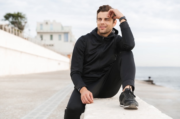 Afbeelding van tevreden sportieve man 30s in zwarte sportkleding, zittend op de promenade aan zee