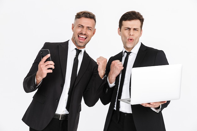Afbeelding van twee gelukkige blanke zakenlieden in kantoorpakken die zich verheugen terwijl ze laptop en mobiele telefoons geïsoleerd houden