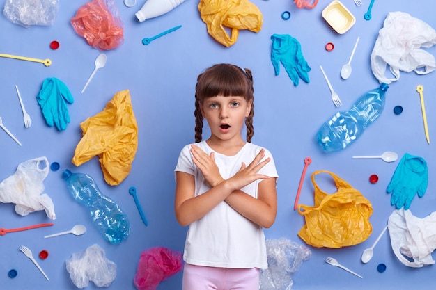 Afbeelding van verbaasd klein meisje in t-shirt kijkend naar camera met open mond die plastic vervuiling laat zien, toont ontkenningsgebaar wil onze planeet redden geplukt plastic afval geïsoleerd op blauwe muur