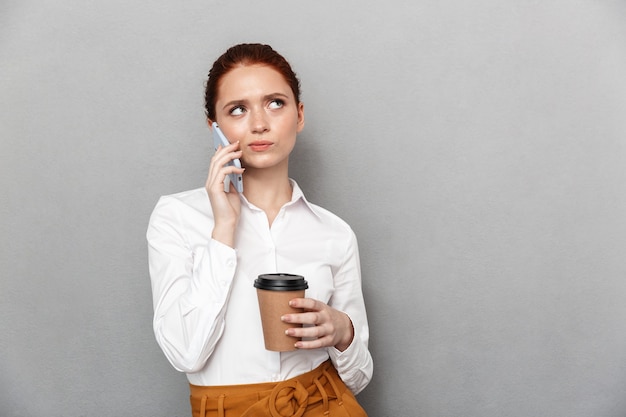 Afbeelding van verwarde jonge mooie roodharige zakenvrouw poseren geïsoleerd over grijze muur praten door mobiele telefoon koffie drinken.