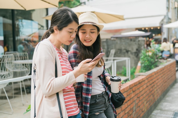 Afbeelding van vrolijke aziatische Koreaanse vrouwelijke reizigers in vrijetijdskleding die afhaalkoffie drinken en smartphone buiten op rode bakstenen muur gebruiken. twee dames genieten van zonneschijn en delen hun mobiele telefoon samen.