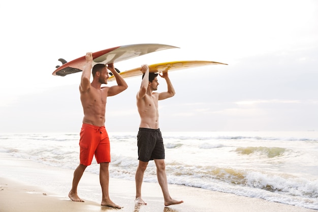 Afbeelding van vrolijke blije jonge twee mannen surfers vrienden met surfen op een strand buiten.