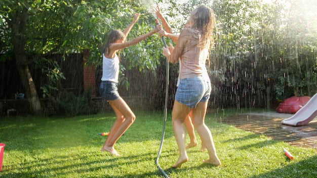 Afbeelding van vrolijke lachende meisjes in natte kleren die dansen in de tuin en een waterslang vasthouden. Familie spelen en plezier hebben buiten in de zomer