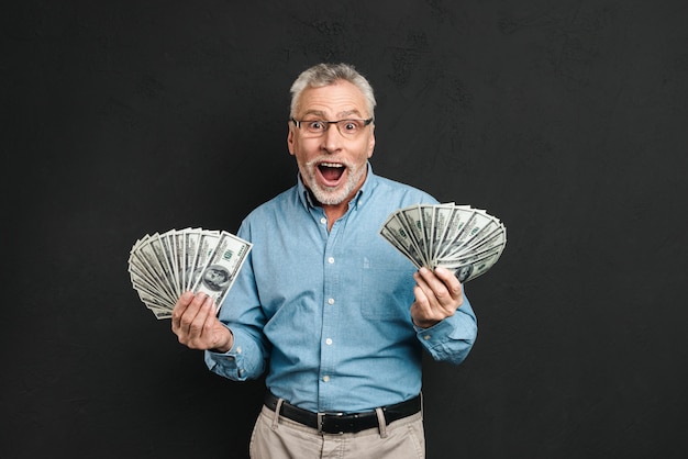 Afbeelding van vrolijke volwassen man jaren 60 met grijs haar met geld twee fans van 100 dollarbiljetten en schreeuwen van geluk, geïsoleerd over zwarte muur