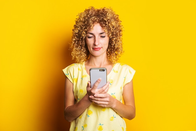 Affable young curly woman looks into the phone on a yellow surface