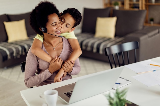 Ragazzo nero affettuoso che bacia sua madre che sta lavorando al computer portatile a casa