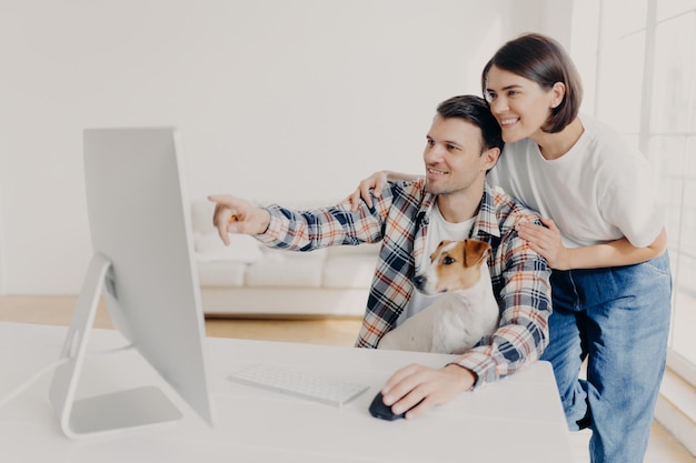 Affectionate brunette wife supports husband with doing project\
work happy man points into monitor tells something shares his ideas\
works together with pedigree dog pose in spacious cabinet