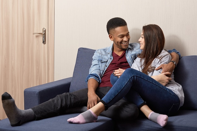 Affectionate mixed race couple in love look happily, have warm hug, dressed casually, pose at sofa in modern apartment