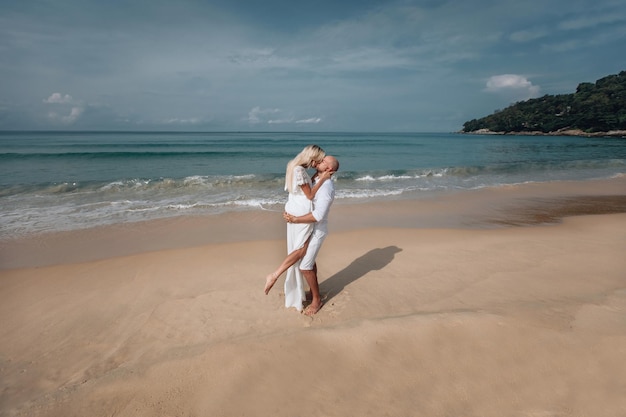 Affectionately embraced and tenderly kissing, a young couple in white clothes enjoys a mid summer  day, on a wet sandy beach. Phuket Thailand.