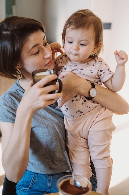 Madre affettuosa che aiuta la sua bambina a bere un bicchiere di latte che tiene sulle sue braccia bella yo