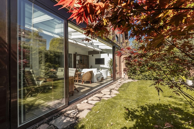 Foto afgesloten terras van bruin aluminium en glas met grote schuifdeuren houten en rieten meubelen binnen een omloop van natuurstenen platen en een fraaie tuin met gazon en sierbomen