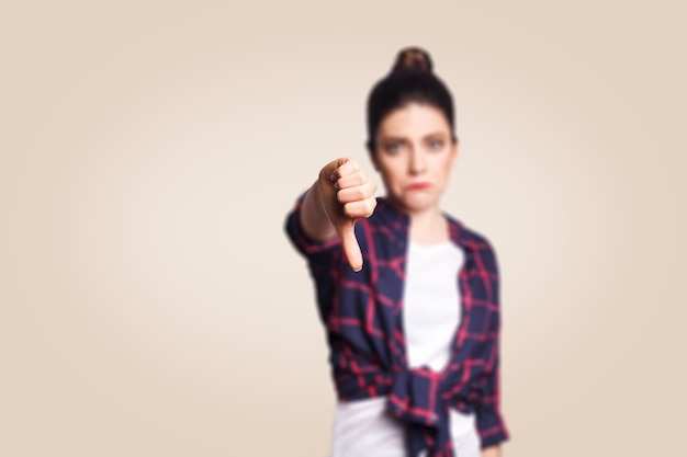 Afkeer. Jong ongelukkig boos meisje met casual stijl en knot haar duimen naar beneden haar vinger, op beige lege muur met kopieerruimte camera kijken met brede glimlach. focus op vinger.