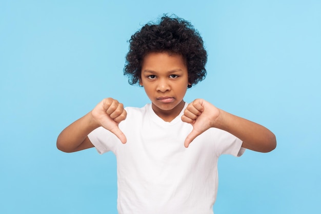 Afkeer. Portret van een ontevreden, overstuur jongetje met krullend haar met een duim omlaag gebaar en een ontevreden, ondeugend kind dat het oneens is met het afkeuren van vingerteken. studio-opname blauwe achtergrond