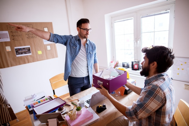 Afraid young employee man sitting on the chair and holding a box with his stuff after getting fired, the unhappy furious boss is pointing away while standing in front of him.