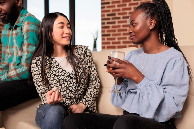 African american adult woman listening, hearing asian friend talking, conversation, drinking wine, eating appetizers at living room friend apartment weekend dinner reunion.