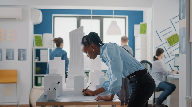 African american architect analyzing blueprints plan and\
building model to create project structure. woman engineer using\
industrial sketch to design architectural construction layout.