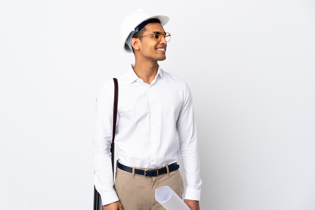 African American architect man with helmet and holding blueprints over isolated white wall _ looking to the side and smiling