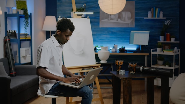 African american artist man searching inspiration on laptop computer sitting in studio space. Black young creative adult working on drawing with online technology for next masterpiece