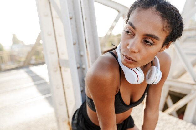 african american beautiful woman with headphones over neck resting while working out on old bridge