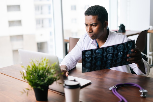 Photo african american black man doctor examining mri brain scan image while working at desk male african neurologist looking xray of patient working on laptop concept of medicine and health care