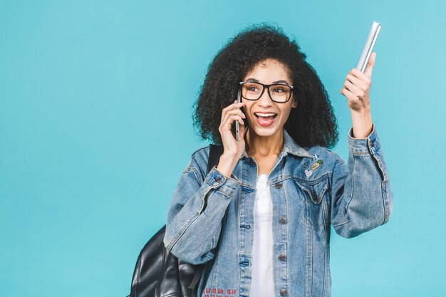 青い背景に分離された上に立っているスマートフォンを使用してアフリカ系アメリカ人の黒い学生女性。