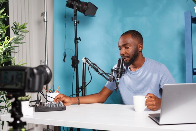 African american blogger creating professional video content with camera and volume controller Vlogger using sound mixer console while live streaming and drinking coffee in studio