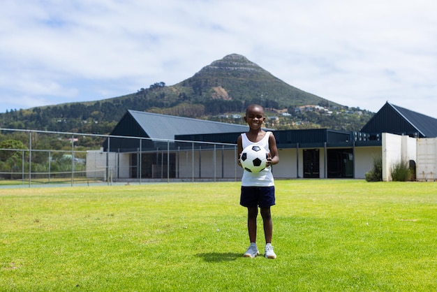 African American boy stands on a grassy field in school holding a soccer ball with copy space