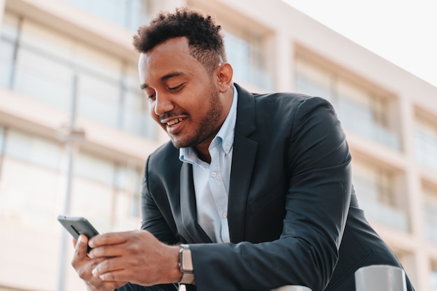Афро-американский мальчик в костюме смотрит в смартфон во время разговора с клиентом своей компании в месте, где видны здания компании. Копировать пространство