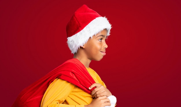 Афро-американский мальчик с Рождеством шляпу и принимая сумку с подарками на красном фоне