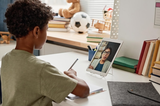 Photo african american boy with curly hair sitting at table in nursery room and making notes while listeni