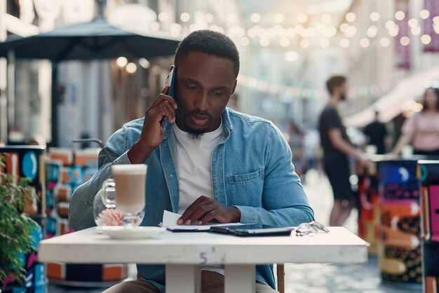 African american business man reads paper documents and works on laptop sitting on outdoor cafe with