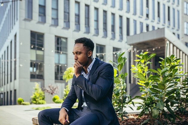 African american business man smoking cannabis outdoors sitting on city park bench on urban street background. Male employee Businessman. Office worker in suit Relieves stress a marijuana outside