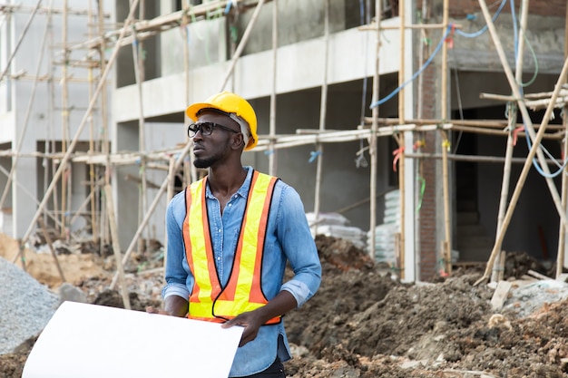 Uomo d'affari afroamericano in elmetto protettivo di sicurezza guardando i piani di blueprint in cantiere.