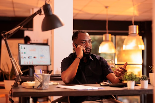 African american businessman talking on mobile phone, having remote conversation with manager in office. Calling number on smartphone, using telephone line to chat about paperwork data.