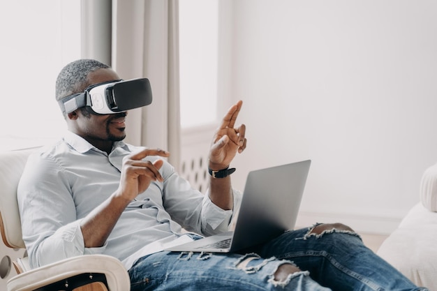 African american businessman in virtual reality glasses working at laptop sitting in armchair