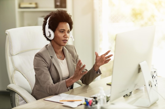 African American businesswoman with headphones having video call while sitting in an office alone at work.