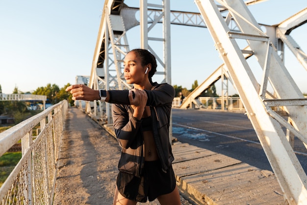 african american charming woman in sportswear using earpods and doing exercises while working out on old bridge