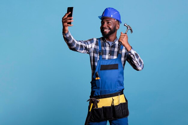 African american contractor taking selfie with mobile phone holding hammer against blue background in studio shot. Construction worker in work uniform and tool belt looking happy.